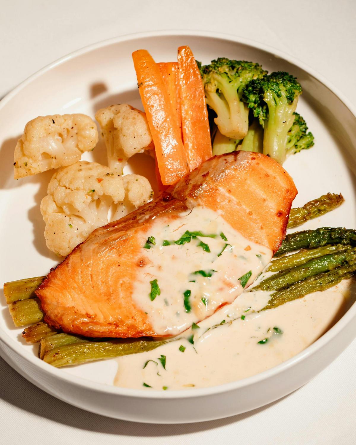 A plate with a grilled salmon fillet topped with creamy herb sauce, served with asparagus, cauliflower, carrots, and broccoli.