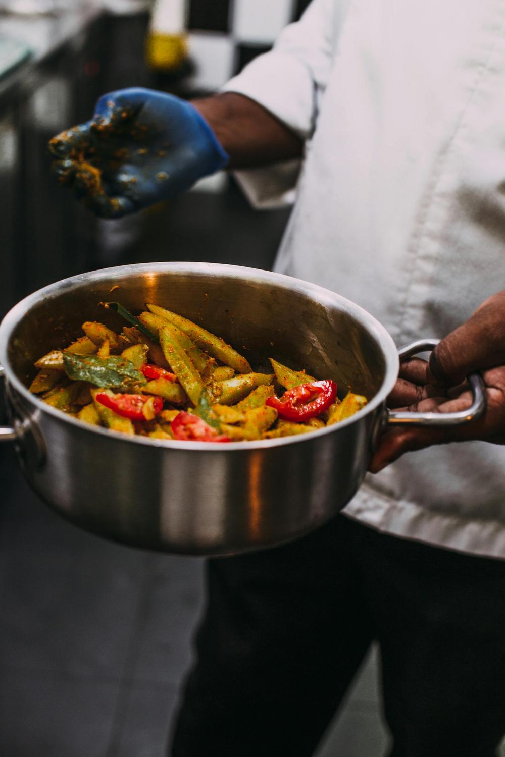 A chef wearing a white coat and blue glove holds a pot filled with seasoned potato wedges and sliced vegetables.