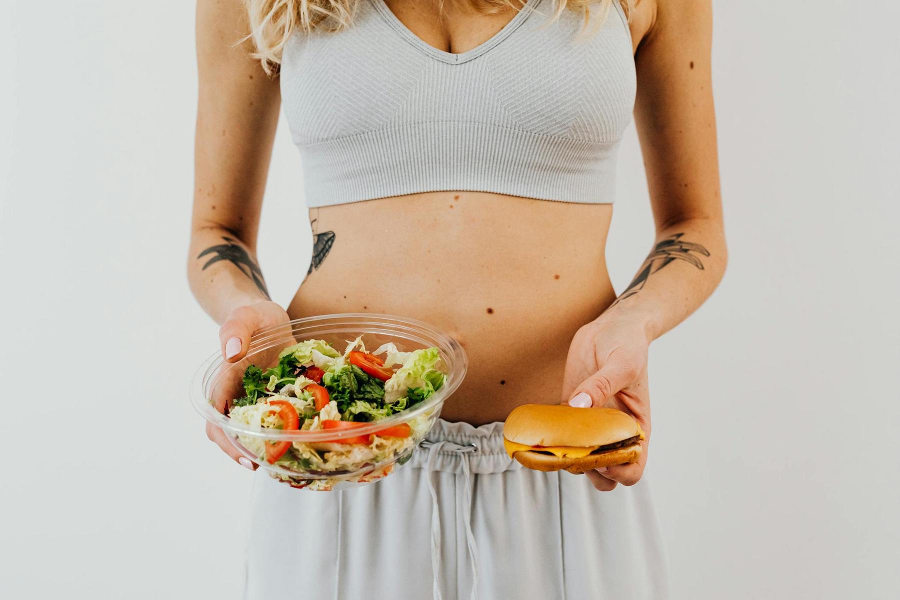 Person in athletic wear holding a salad in one hand and a burger in the other, standing against a plain background.