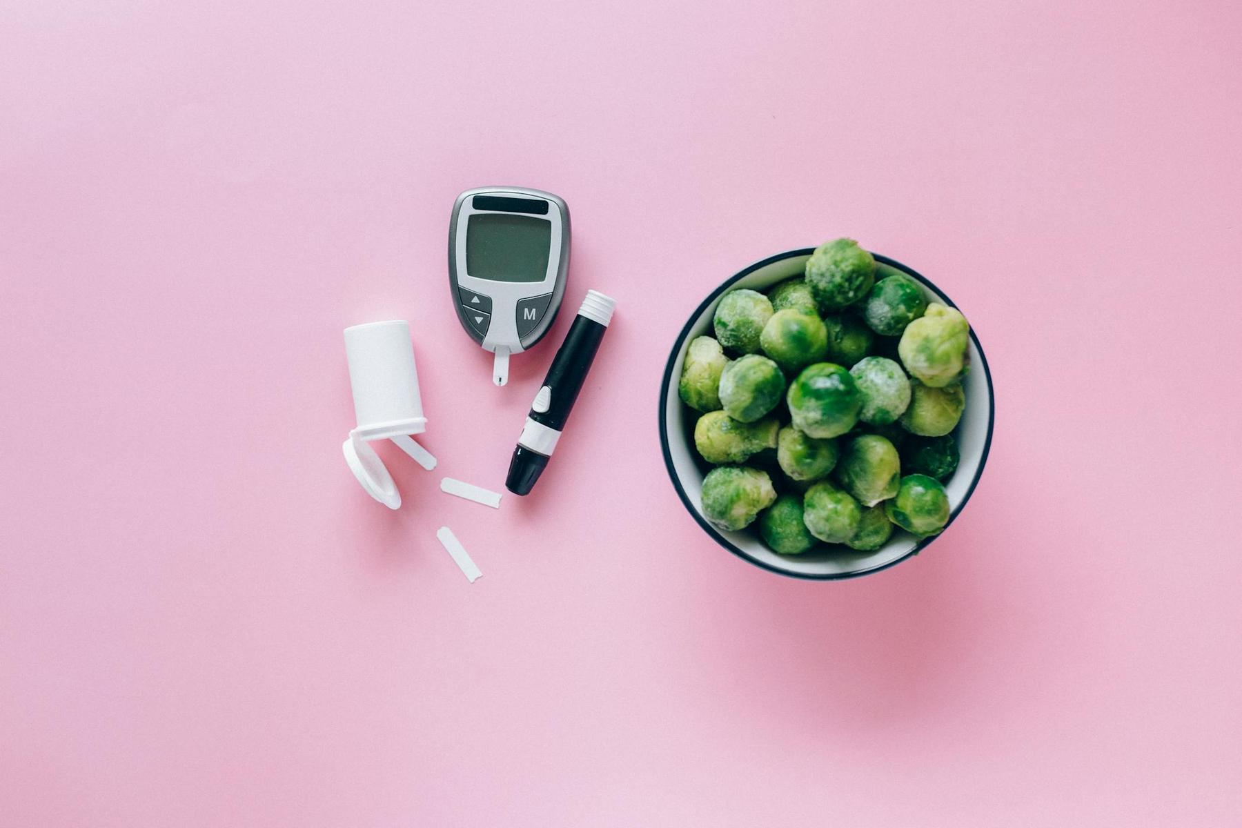 A bowl of Brussels sprouts, a glucose meter, test strips, and a white lancing device on a pink background.