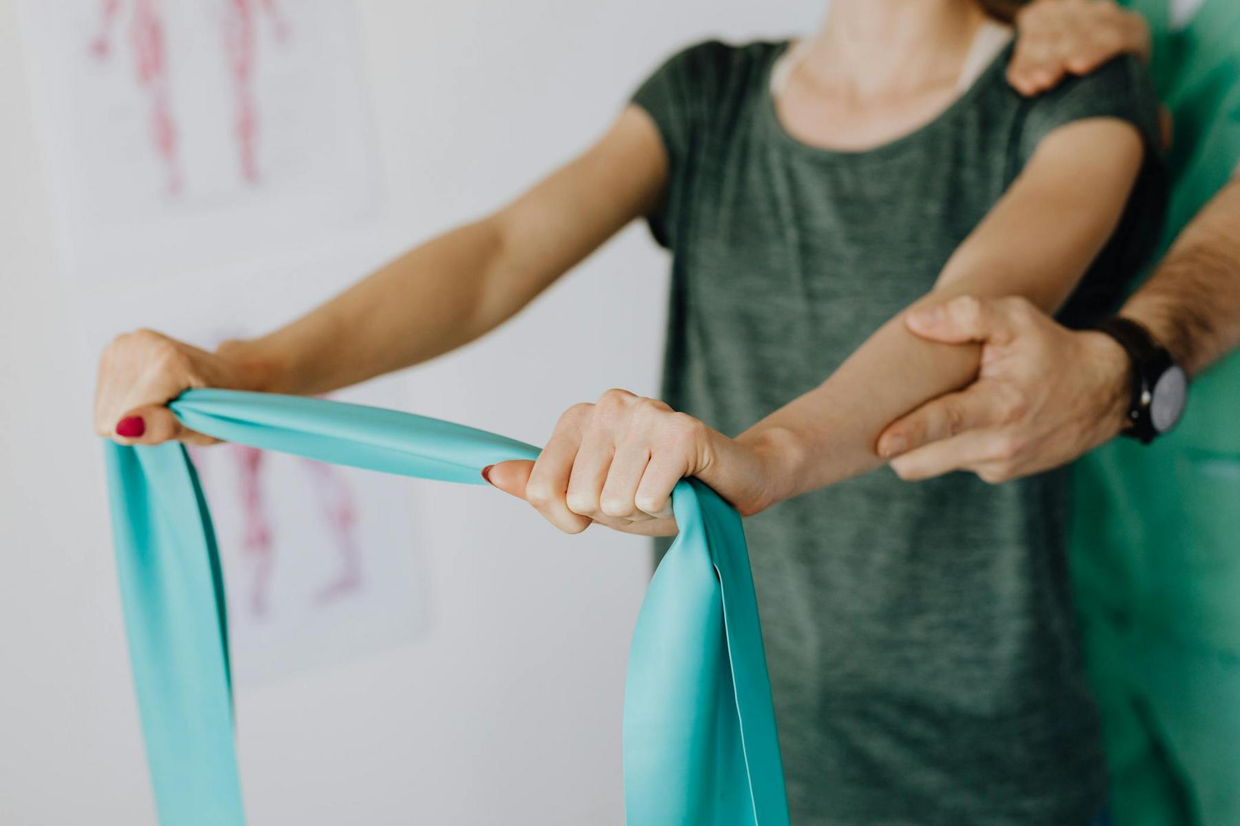 A person in a green shirt stretches an exercise band while another person supports their arm, likely during a physical therapy session.