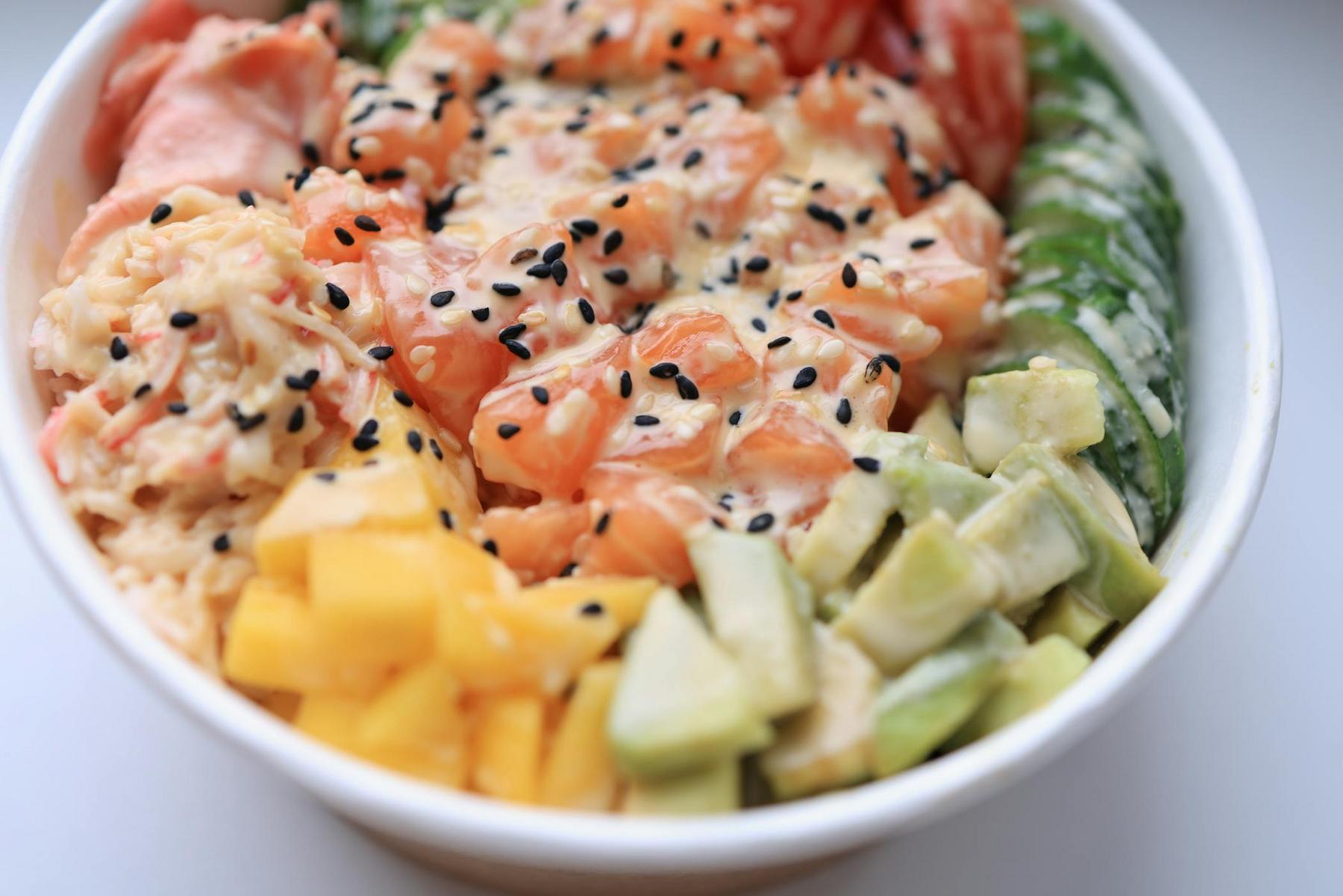 A poke bowl with diced salmon, avocado, mango, cucumber, and rice, topped with black sesame seeds and a drizzle of sauce.