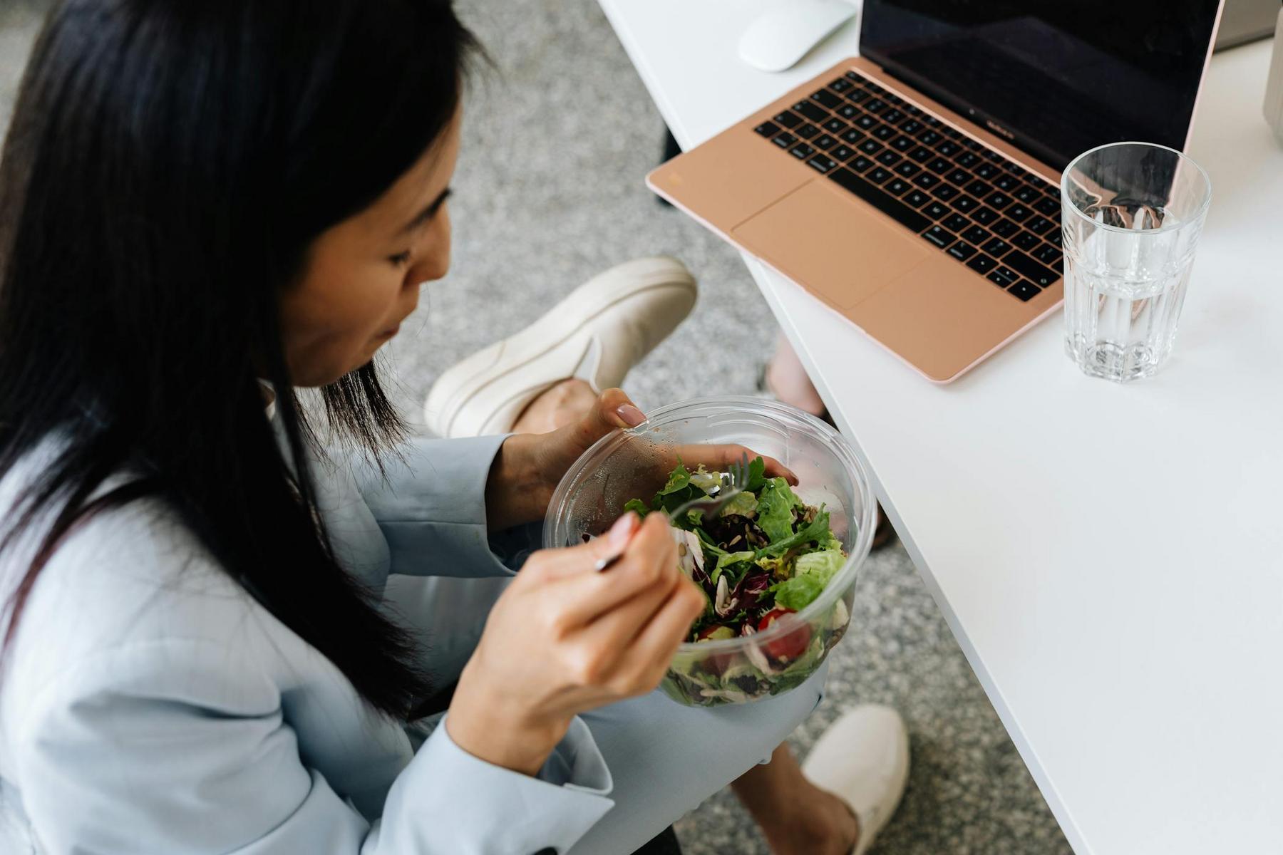 A person in a light suit is sitting at a table, eating a salad from a clear bowl. A laptop and a glass of water are on the table next to them.