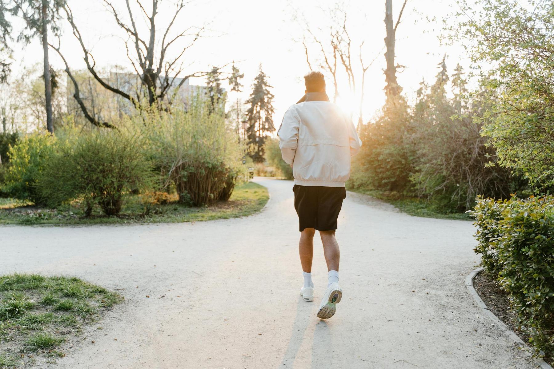 A person wearing a light jacket and shorts jogs on a gravel path in a park with greenery on both sides and the sun shining through the trees.