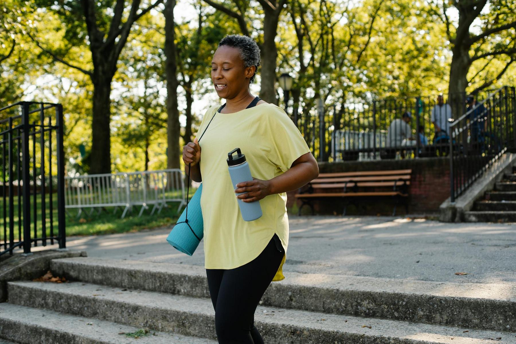 A person in workout clothes walks outdoors holding a water bottle and a rolled-up yoga mat, with trees and benches in the background.