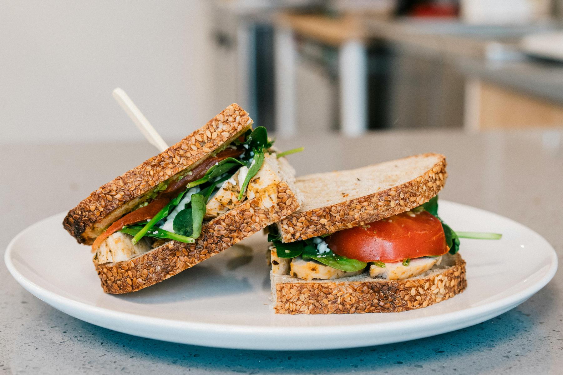 Two whole grain sandwiches with chicken, spinach, and tomato slices are placed on a white plate on a kitchen counter.