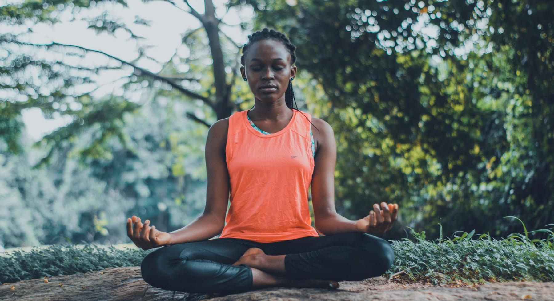 A person sits cross-legged on the ground outdoors, eyes closed, with hands resting on knees, practicing meditation. Trees and greenery are visible in the background.
