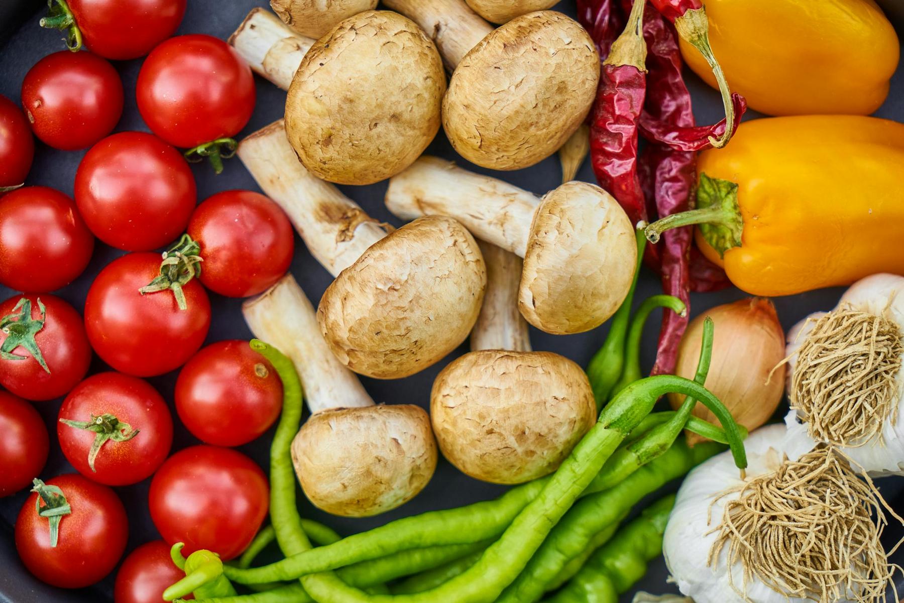 Assorted vegetables including cherry tomatoes, mushrooms, yellow bell peppers, green chili peppers, dried red chilies, garlic, and shallots laid out on a surface.