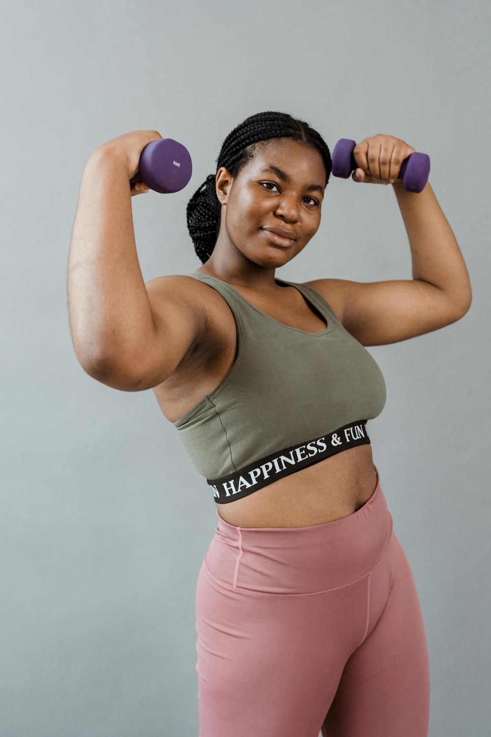 A woman wearing a green sports bra and pink leggings holds two purple dumbbells, flexing her arms and looking at the camera against a plain gray background.