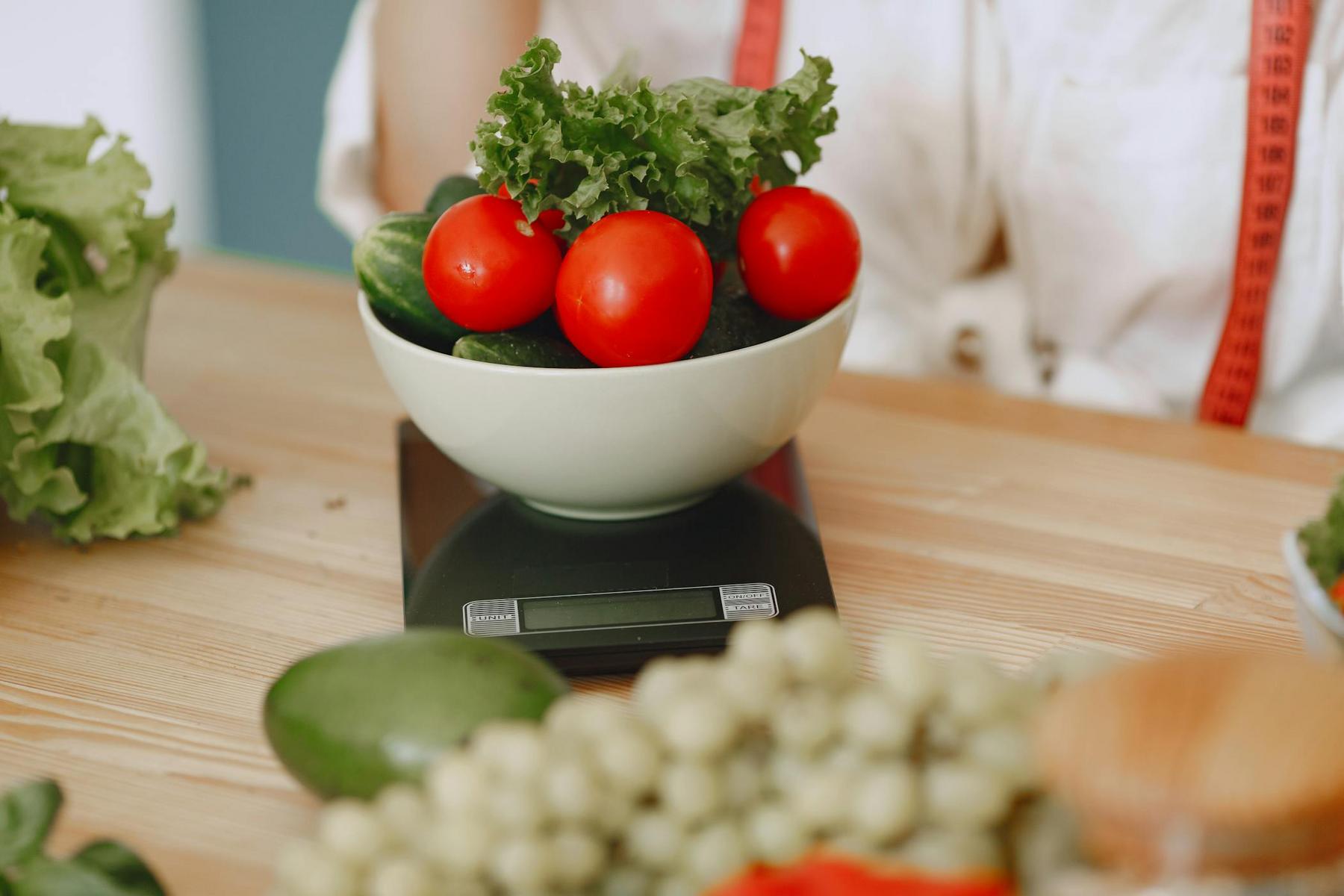 A bowl with lettuce, tomatoes, and cucumbers sits on a digital kitchen scale, surrounded by other fruits and vegetables on a wooden table.