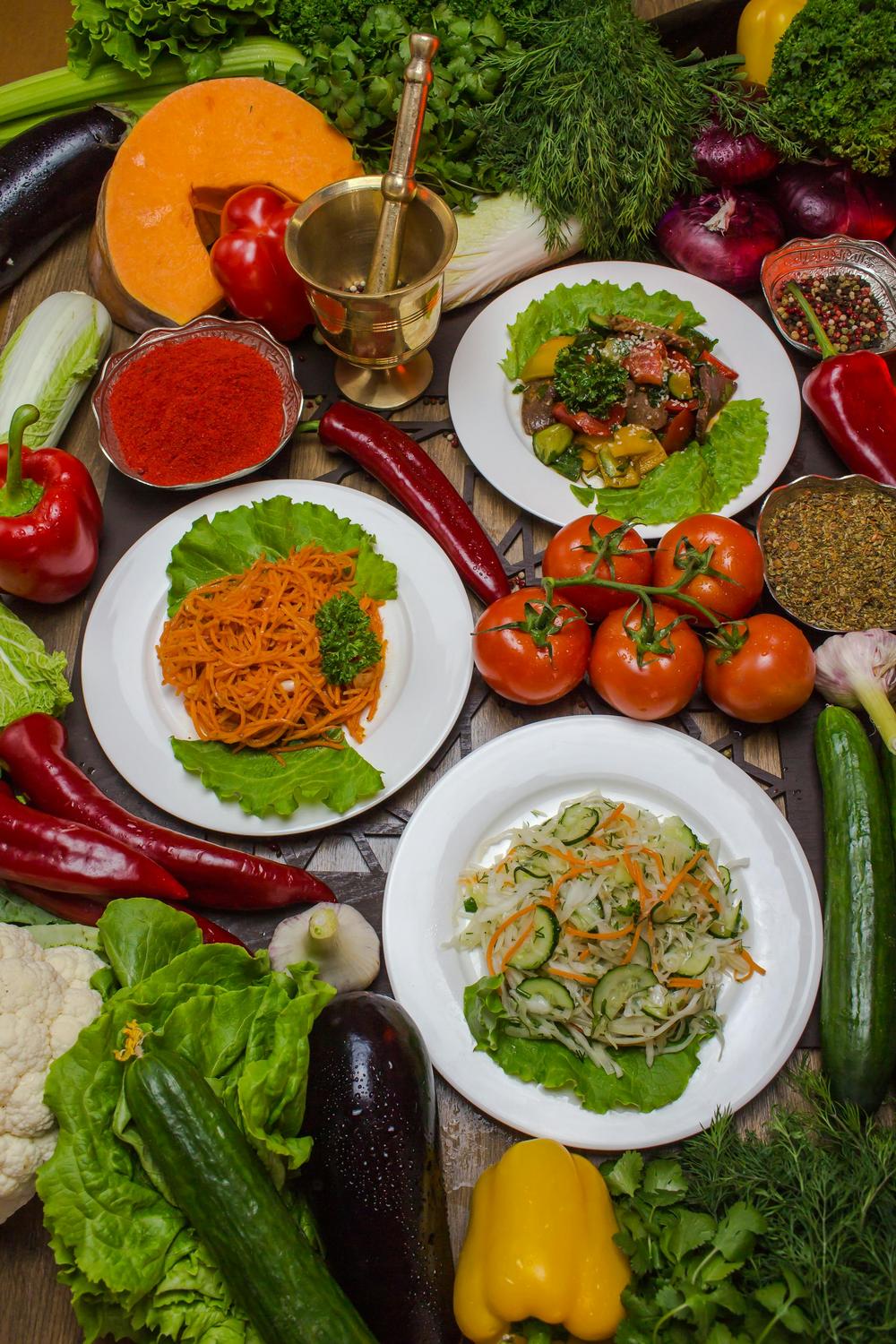 A variety of fresh vegetables and herbs surround three plates of salads and a mortar and pestle on a wooden surface.