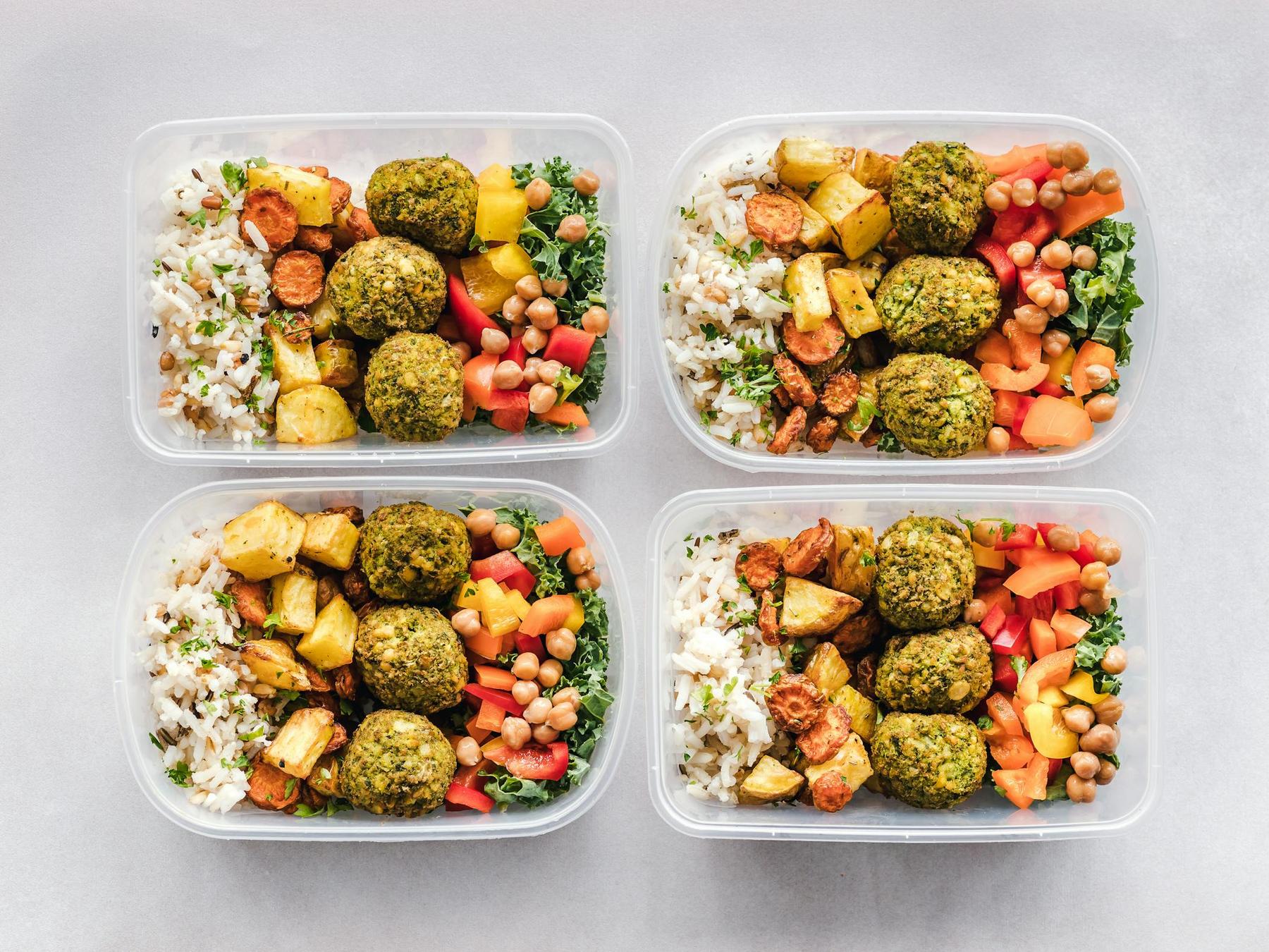 Four meal prep containers each filled with rice, falafel, roasted potatoes, chickpeas, chopped tomatoes, and greens, arranged on a light surface.