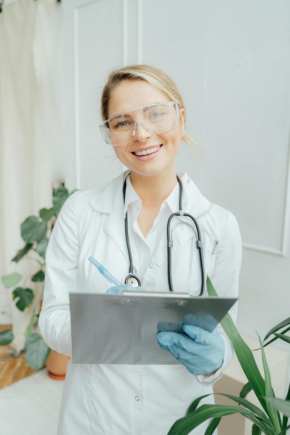 A healthcare professional wearing a lab coat, stethoscope, gloves, and safety glasses smiles while writing on a clipboard in a bright room with plants.
