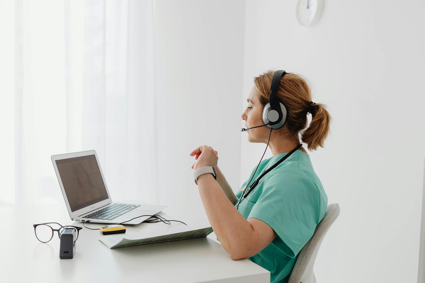 A healthcare worker in scrubs and a headset sits at a desk, looking at a laptop screen, with a notepad, glasses, and medical device nearby.