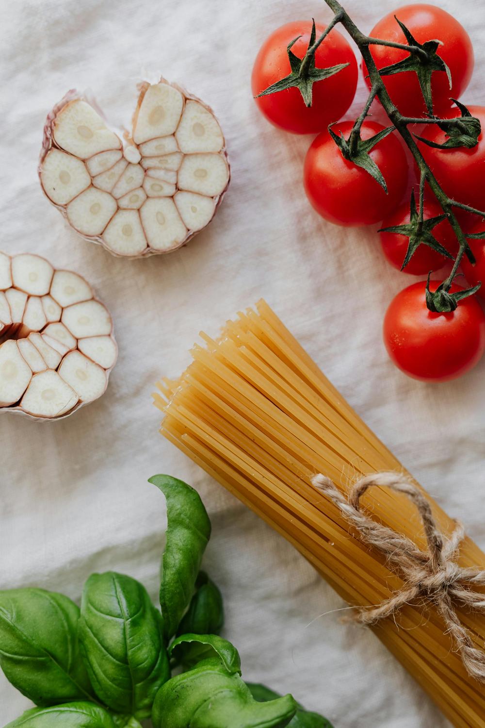 Uncooked spaghetti, tomatoes on the vine, halved garlic bulbs, and fresh basil leaves arranged on a white cloth background.