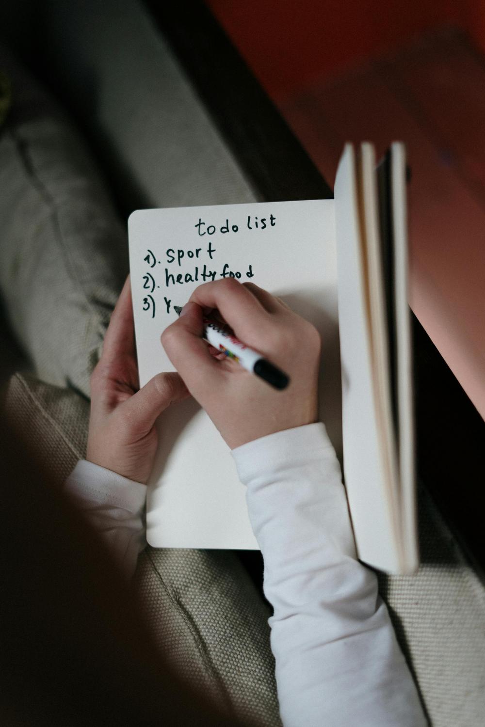 A person writes a to-do list in a notebook with three items: sport, healthy food, and an unfinished third item.