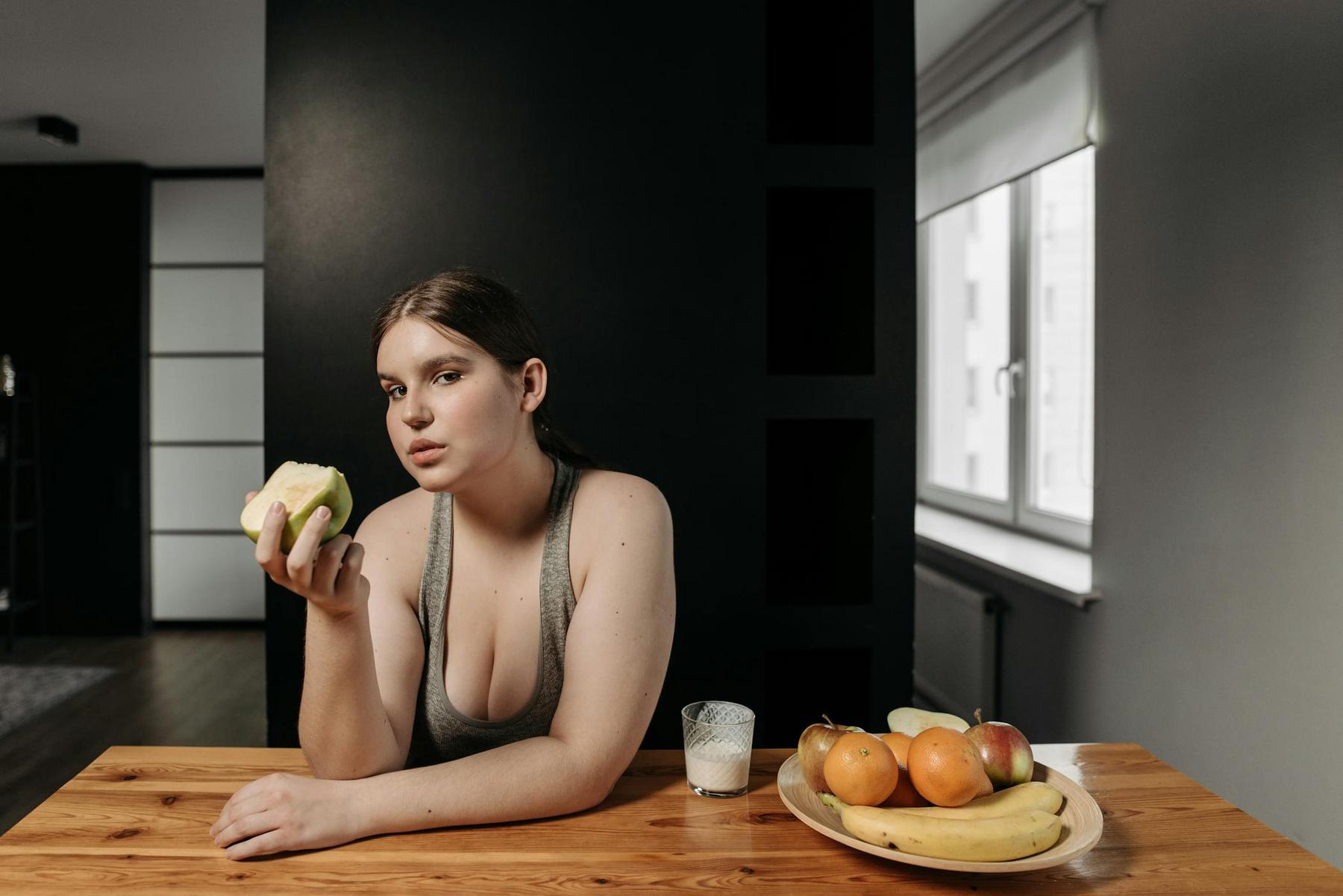A woman sits at a wooden table holding a green apple, with a glass of milk and a plate of assorted fruit in front of her.