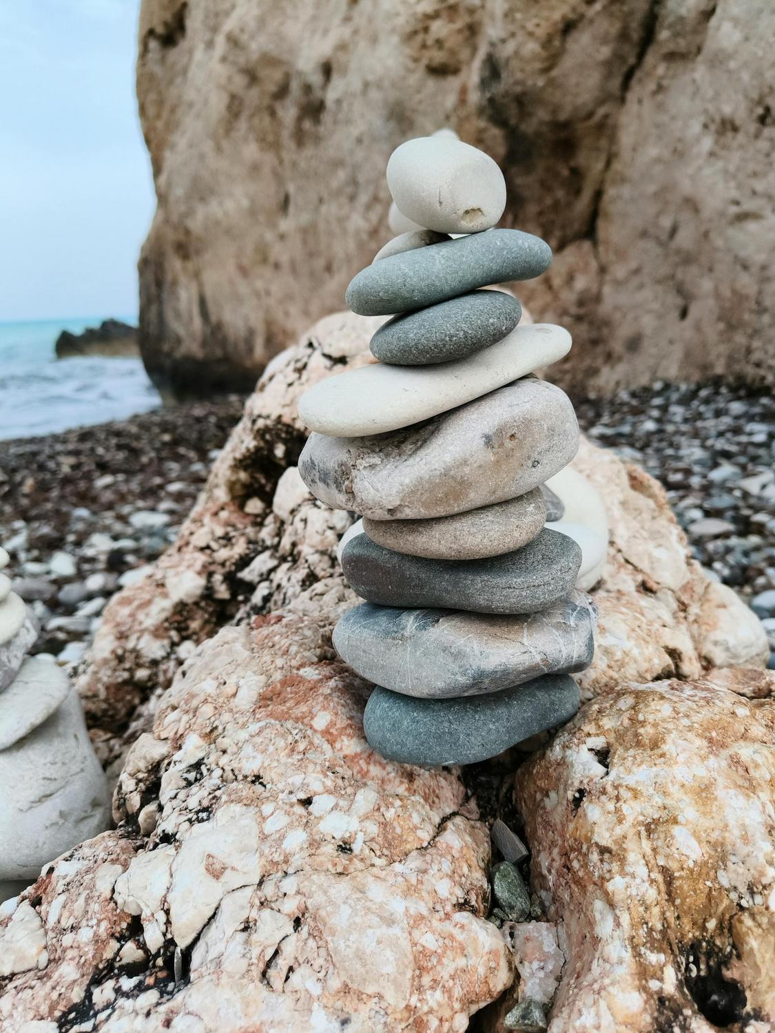 A stack of smooth, flat stones is balanced on a large rock by a rocky beach, with the sea and a cliff visible in the background.