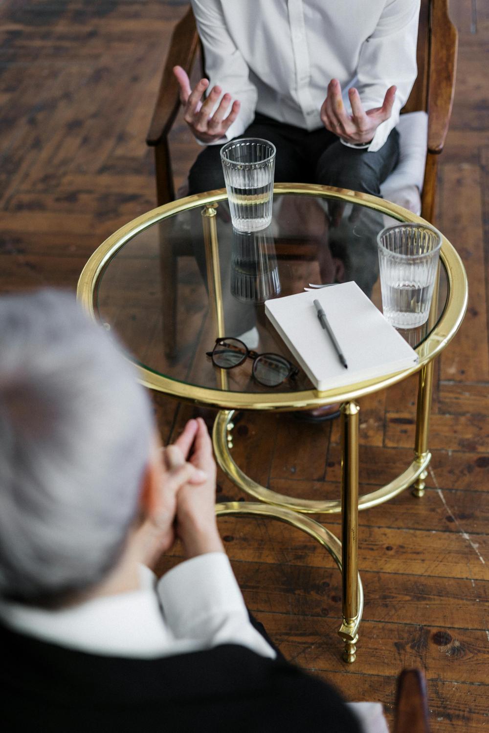 Two people sit across from each other at a round glass table with two glasses of water, a notebook, a pen, and a pair of eyeglasses on it.