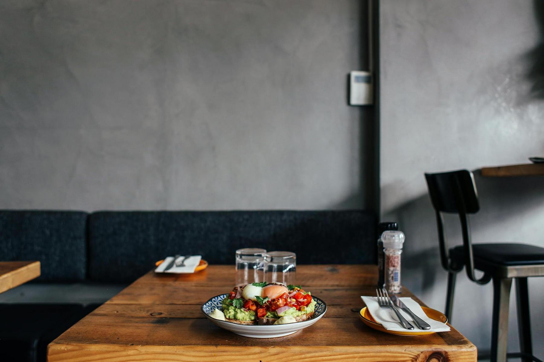 A wooden table in a restaurant with a plate of food, two glasses of water, utensils, and a pepper grinder, with empty chairs nearby.