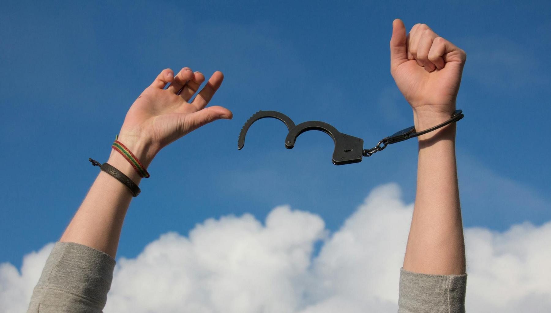 Two raised hands, one wearing an open handcuff, are shown against a blue sky with clouds.