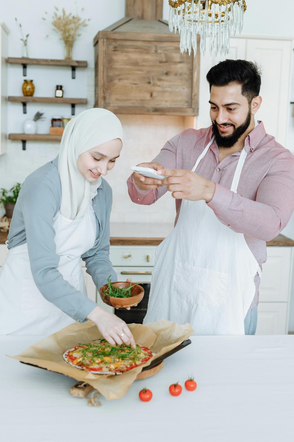 Two people in aprons prepare a pizza in a kitchen; one adds greens to the pizza while the other takes a photo with a smartphone.