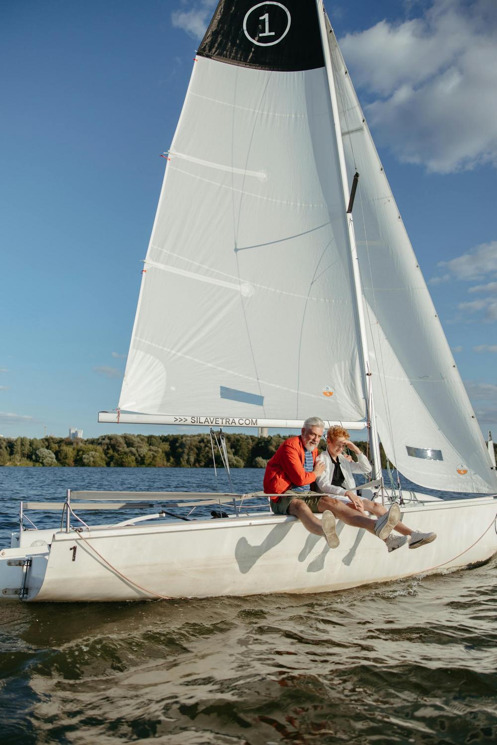 Two people sit on the edge of a white sailboat with a large sail, sailing on a body of water under a blue sky with clouds.