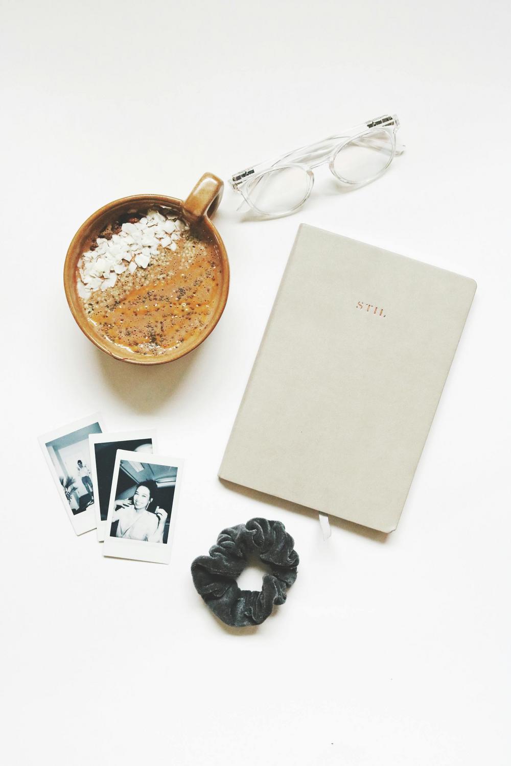 A cup of oatmeal, clear eyeglasses, a beige notebook labeled "STIL," a black scrunchie, and three instant photos are arranged on a white surface.