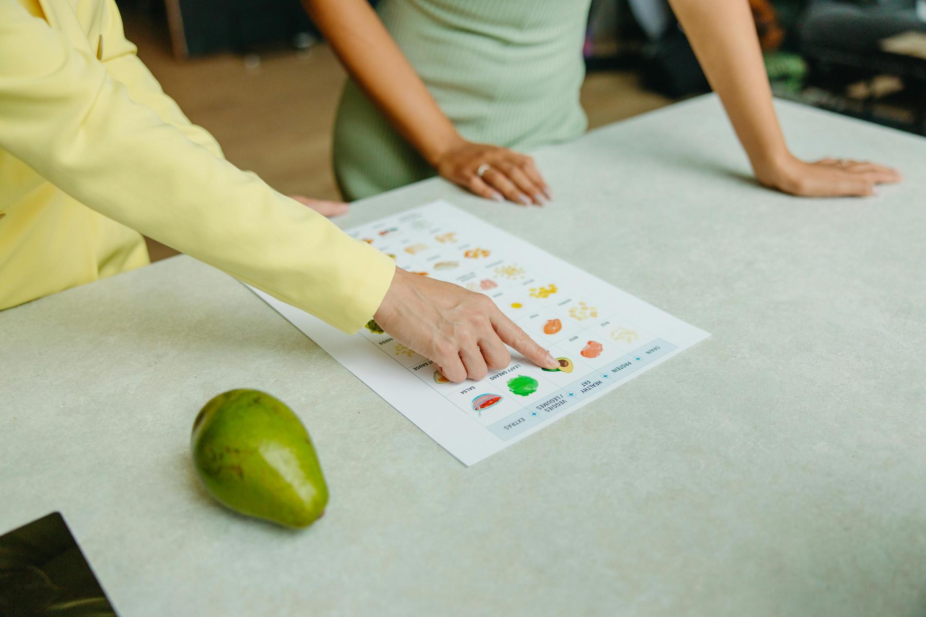 Two people stand at a table, one pointing to a food chart with various images and text, while an avocado rests on the tabletop.