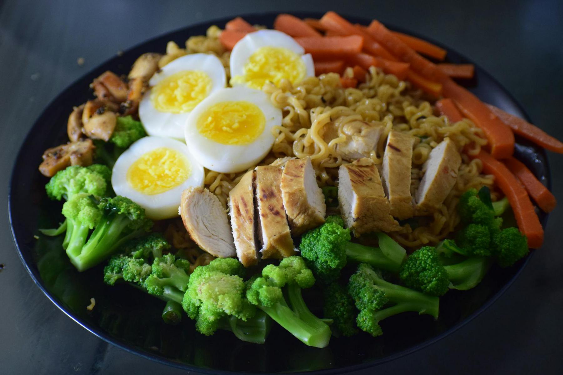 A black plate with boiled eggs, sliced grilled chicken, broccoli, carrot sticks, and cooked noodles arranged in separate sections.