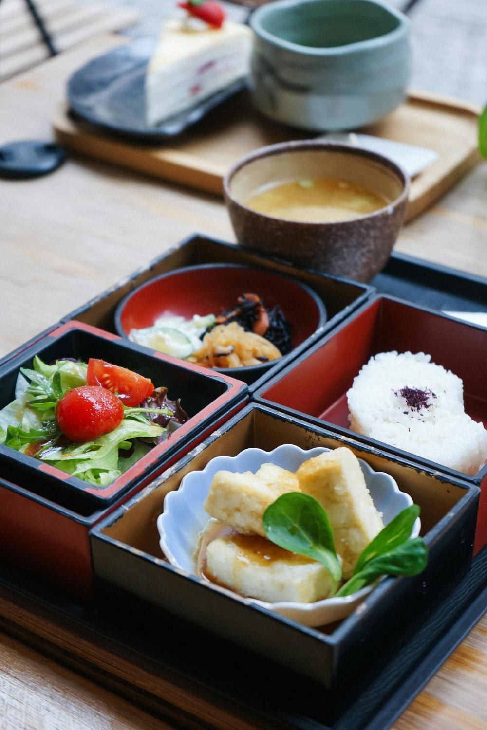 A Japanese bento box with rice, salad, tofu, vegetables, and a bowl of soup, served on a wooden table with a tea cup and dessert in the background.