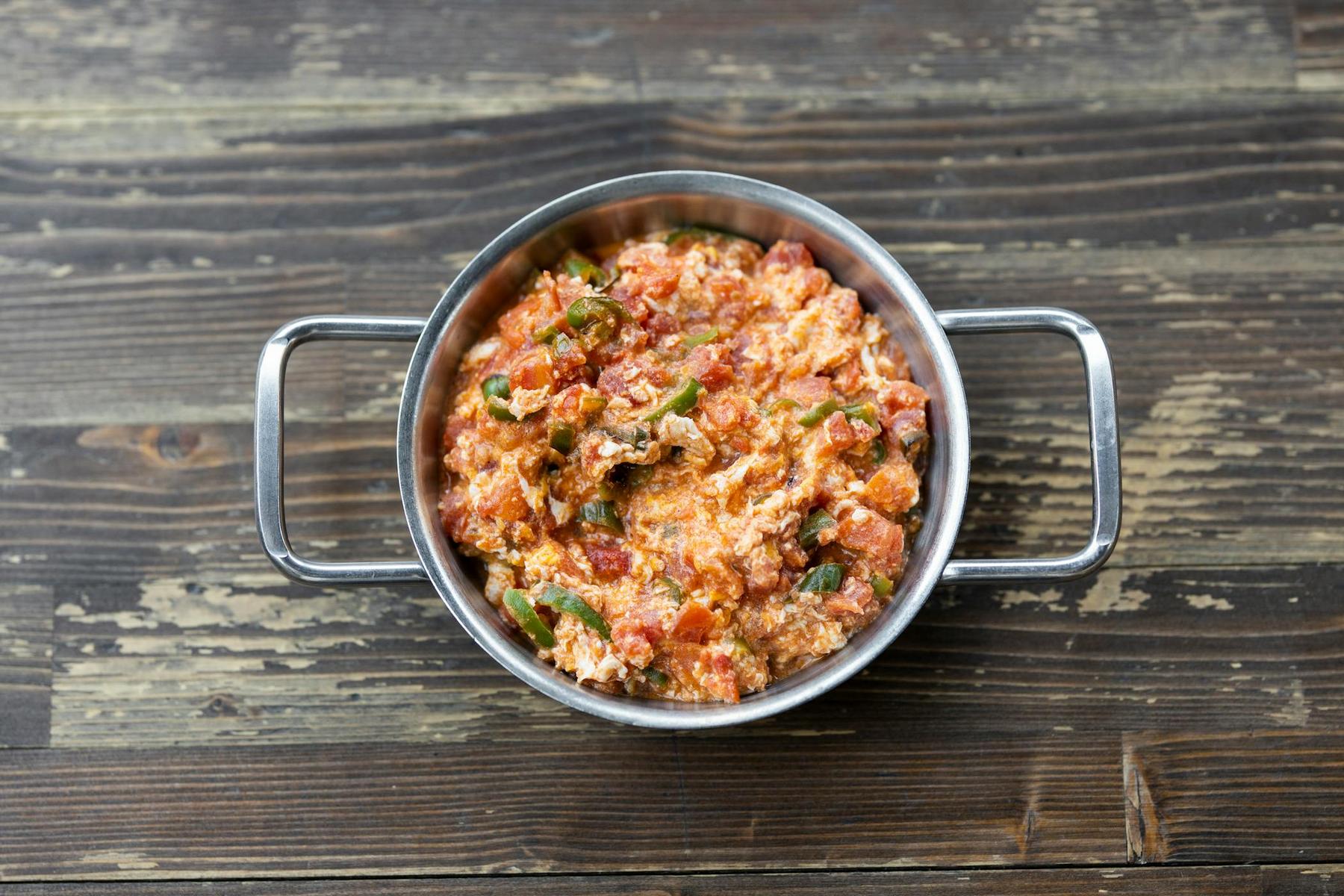 A metal bowl with handles contains a tomato-based scrambled egg dish with green peppers, set on a wooden surface.