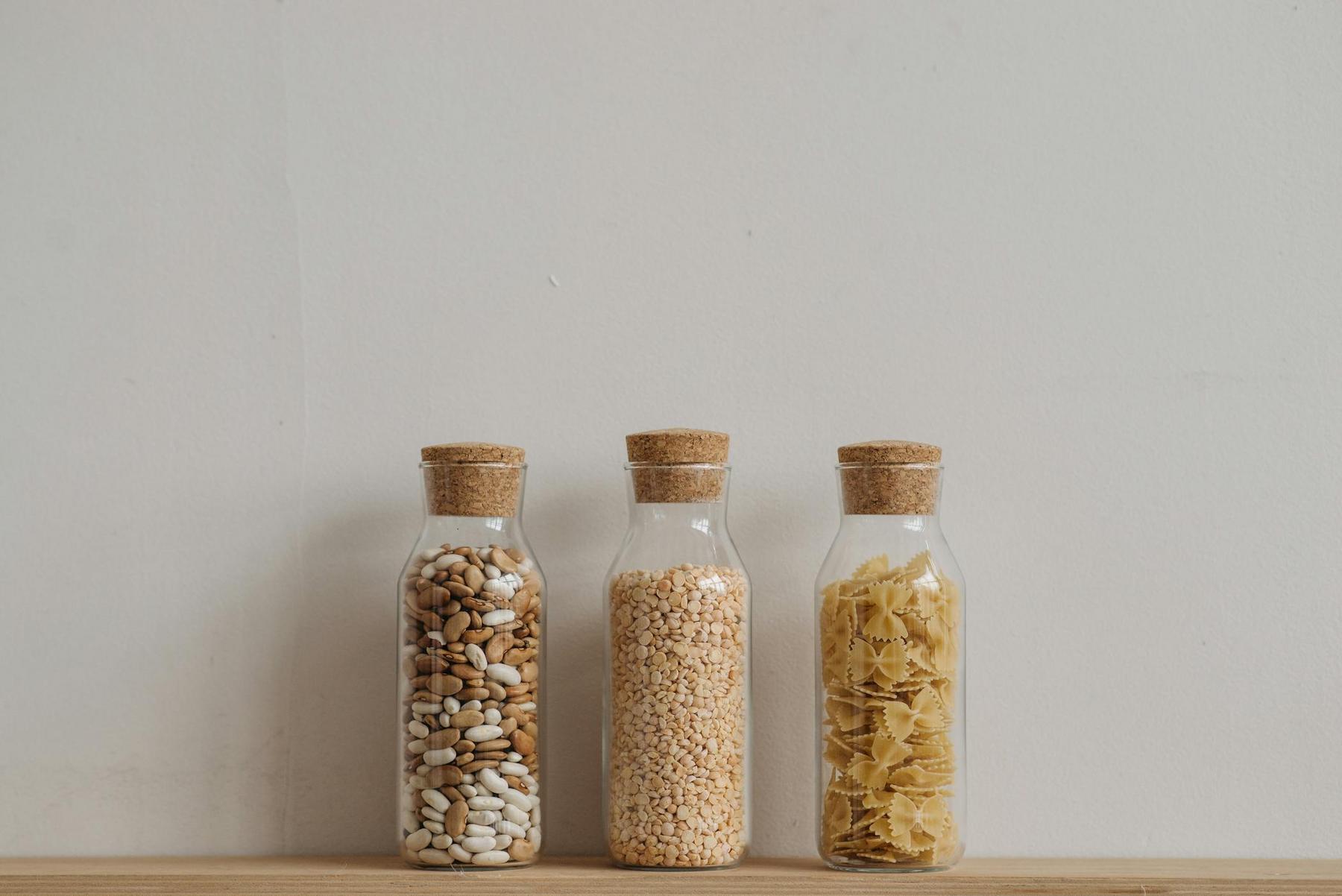 Three glass jars with cork lids, containing dried beans, lentils, and pasta, are placed in a row on a wooden surface against a plain white wall.