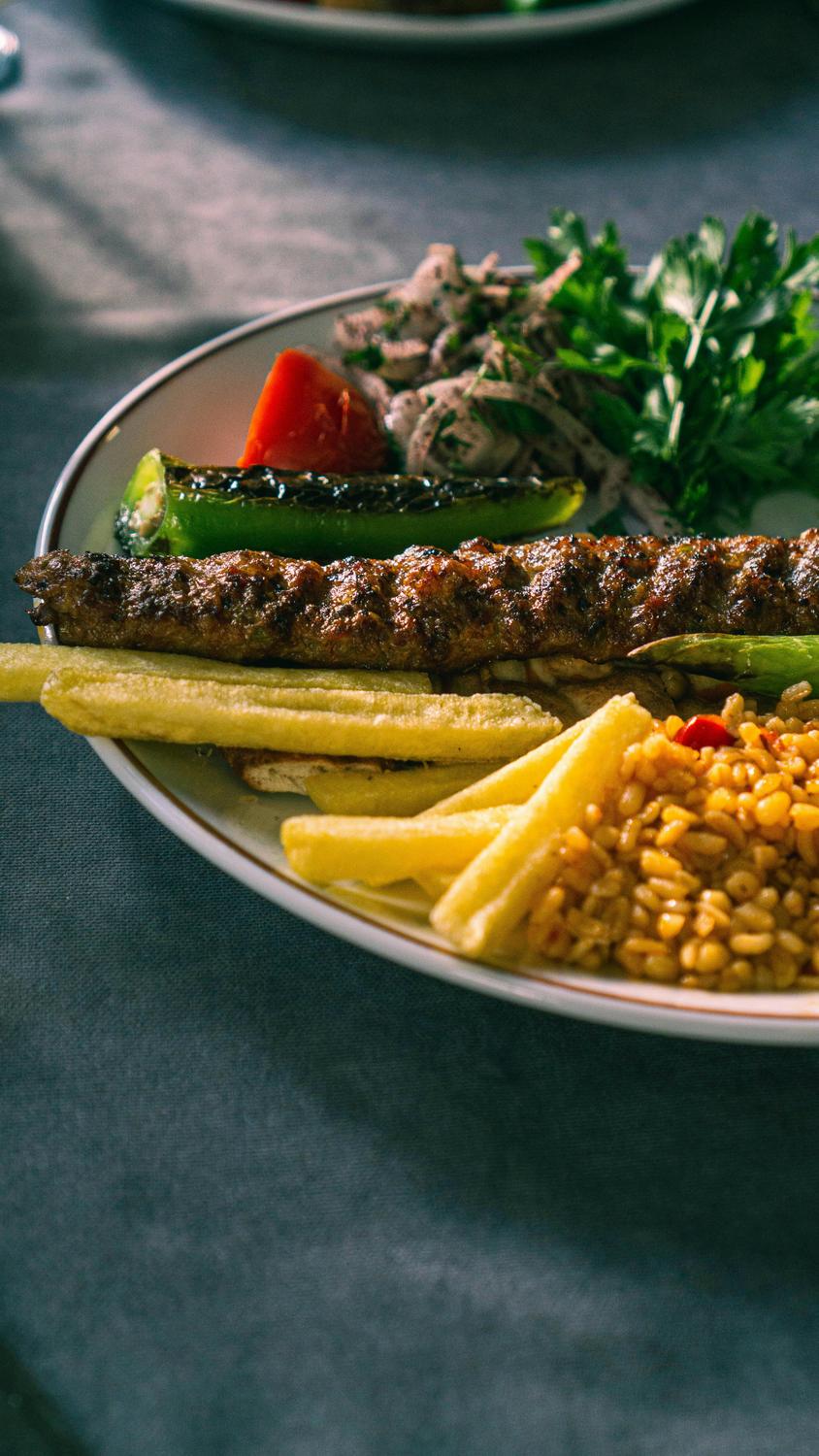 A plate with grilled kebab, French fries, bulgur, grilled vegetables, fresh herbs, and onion salad on a table.