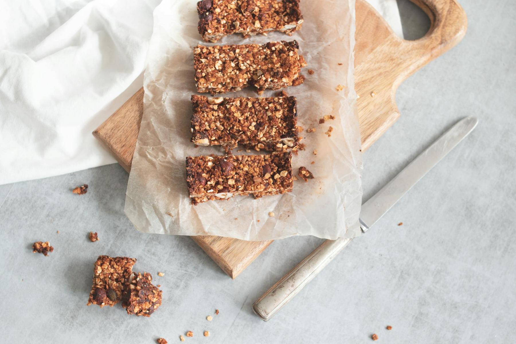Four granola bars are arranged on parchment paper atop a wooden cutting board, with a knife beside them and some crumbs scattered on a gray surface.