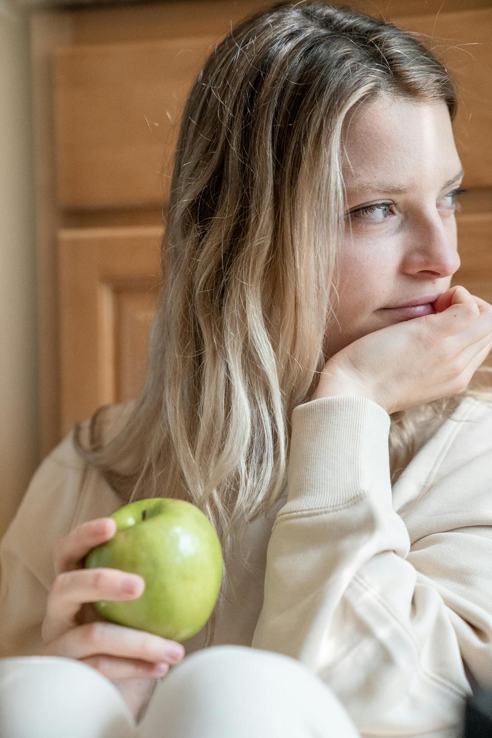 A woman with long blonde hair holds a green apple and looks pensively to the side, resting her chin on her hand.
