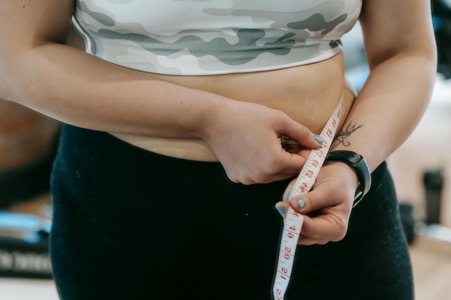 A person measuring their waist with a tape measure, wearing a camouflage sports bra and black leggings.
