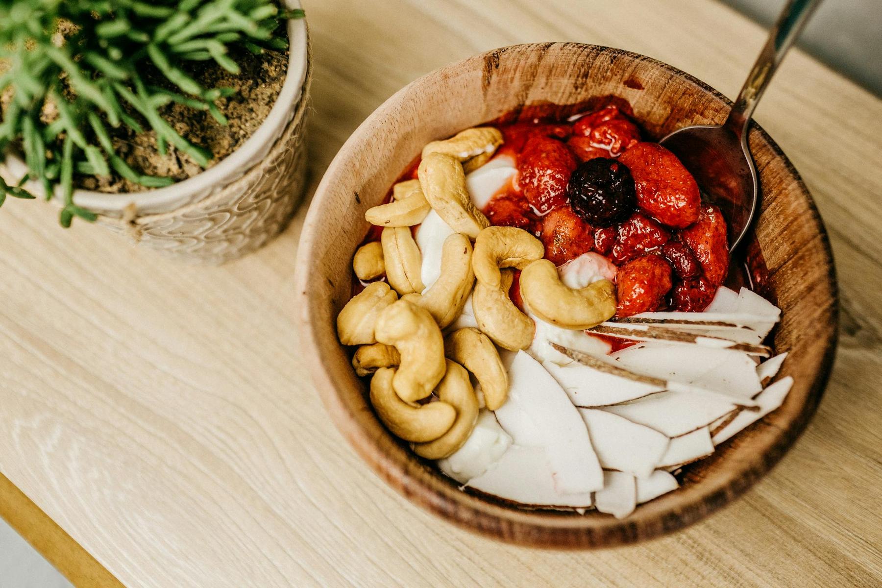 A wooden bowl on a table contains cashews, strawberries, cherries, and coconut slices. A spoon rests inside. A small potted plant is next to the bowl.