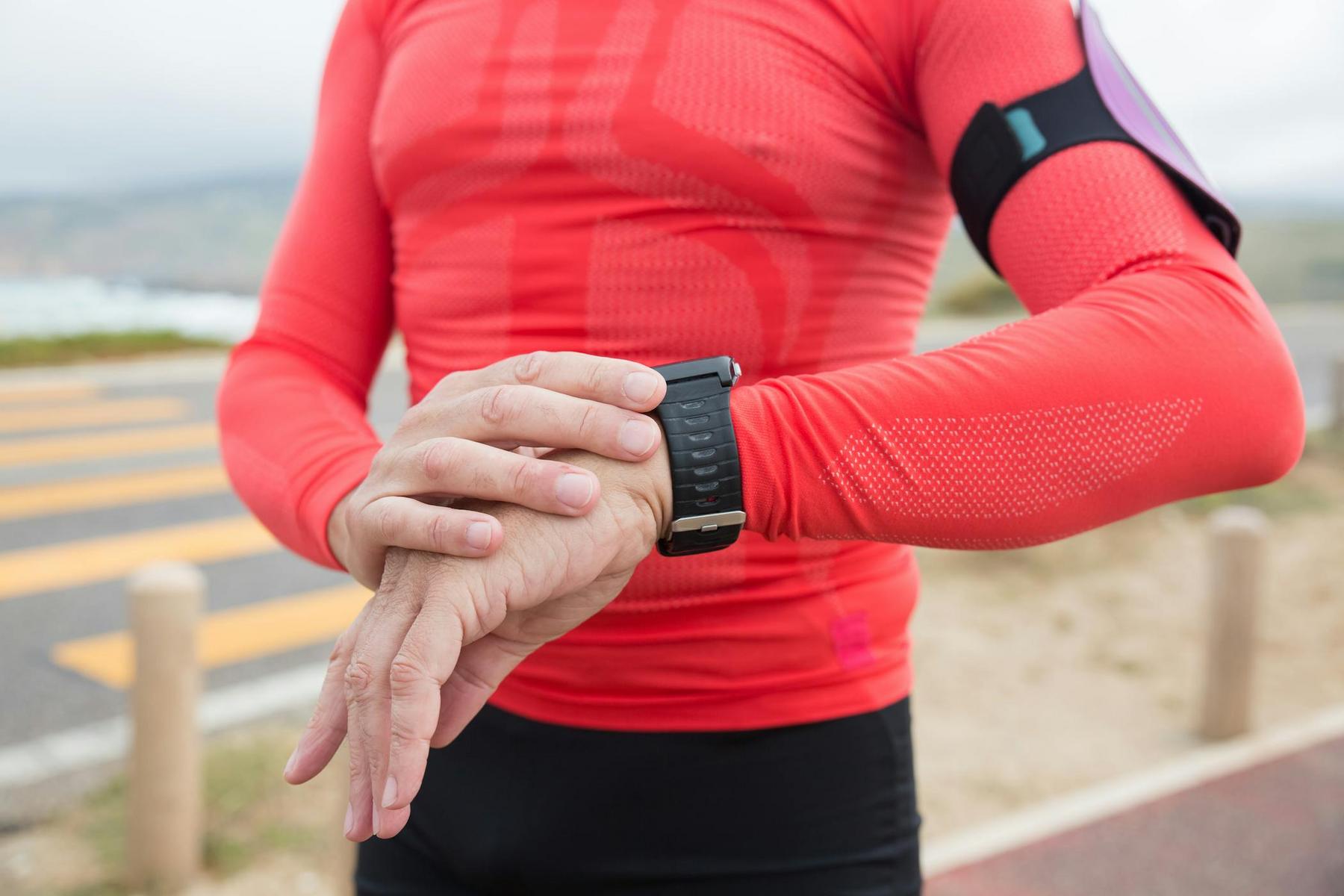 A person in a red long-sleeve athletic shirt checks a smartwatch outdoors near a road with crosswalk markings.