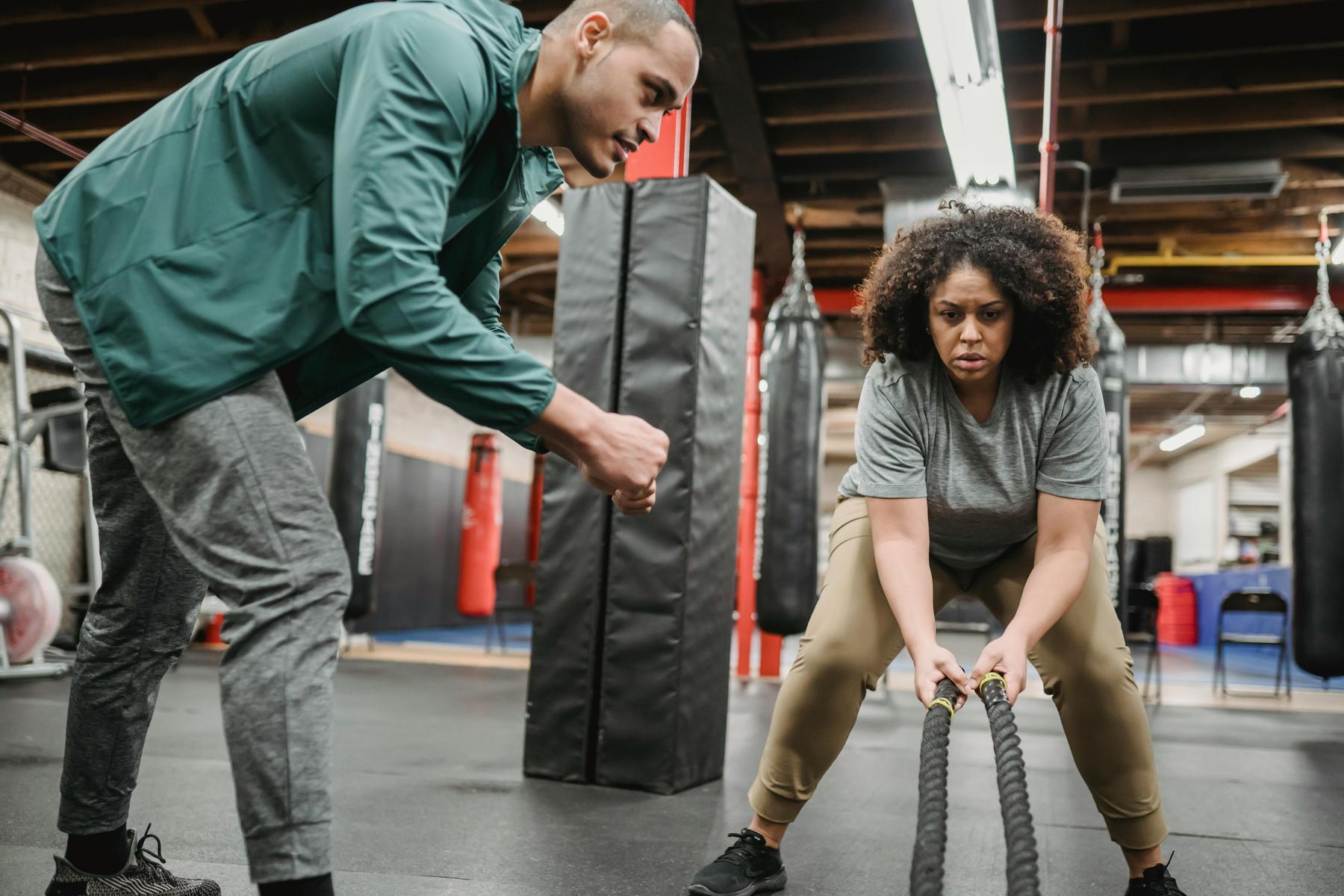 A woman exercises with battle ropes in a gym while a trainer observes and encourages her.