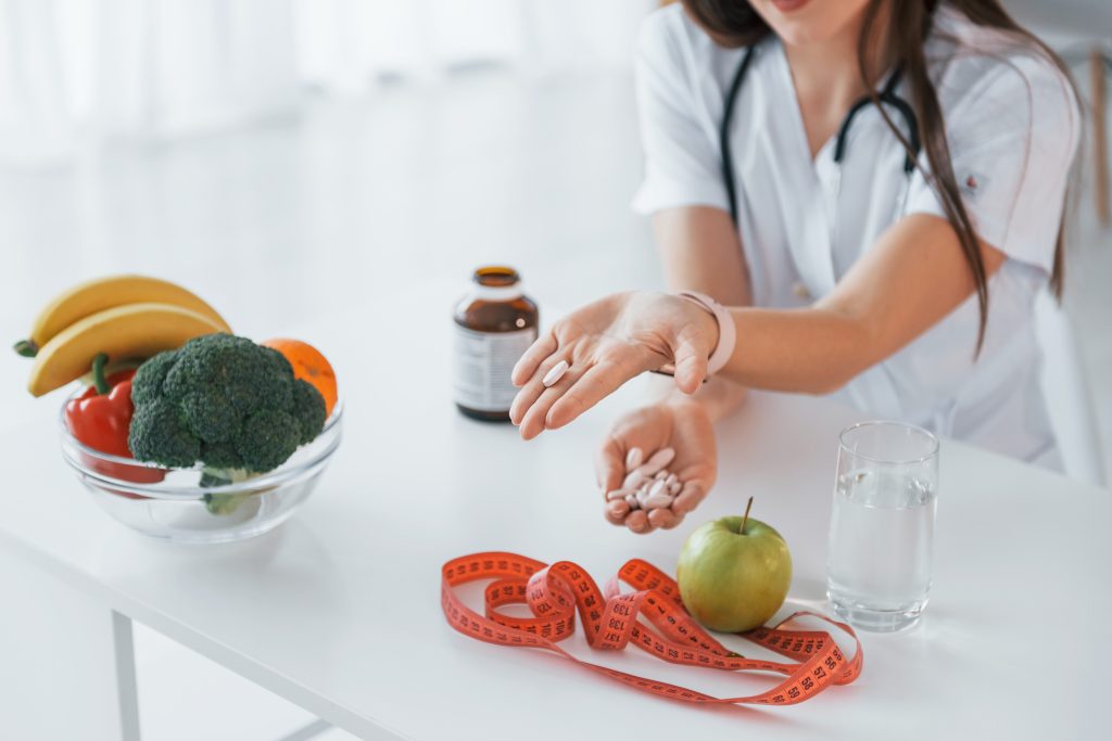 A person in a white coat sits at a table with fruits, vegetables, a measuring tape, a bottle, and a glass of water, exploring the science of appetite while holding pills in one hand.