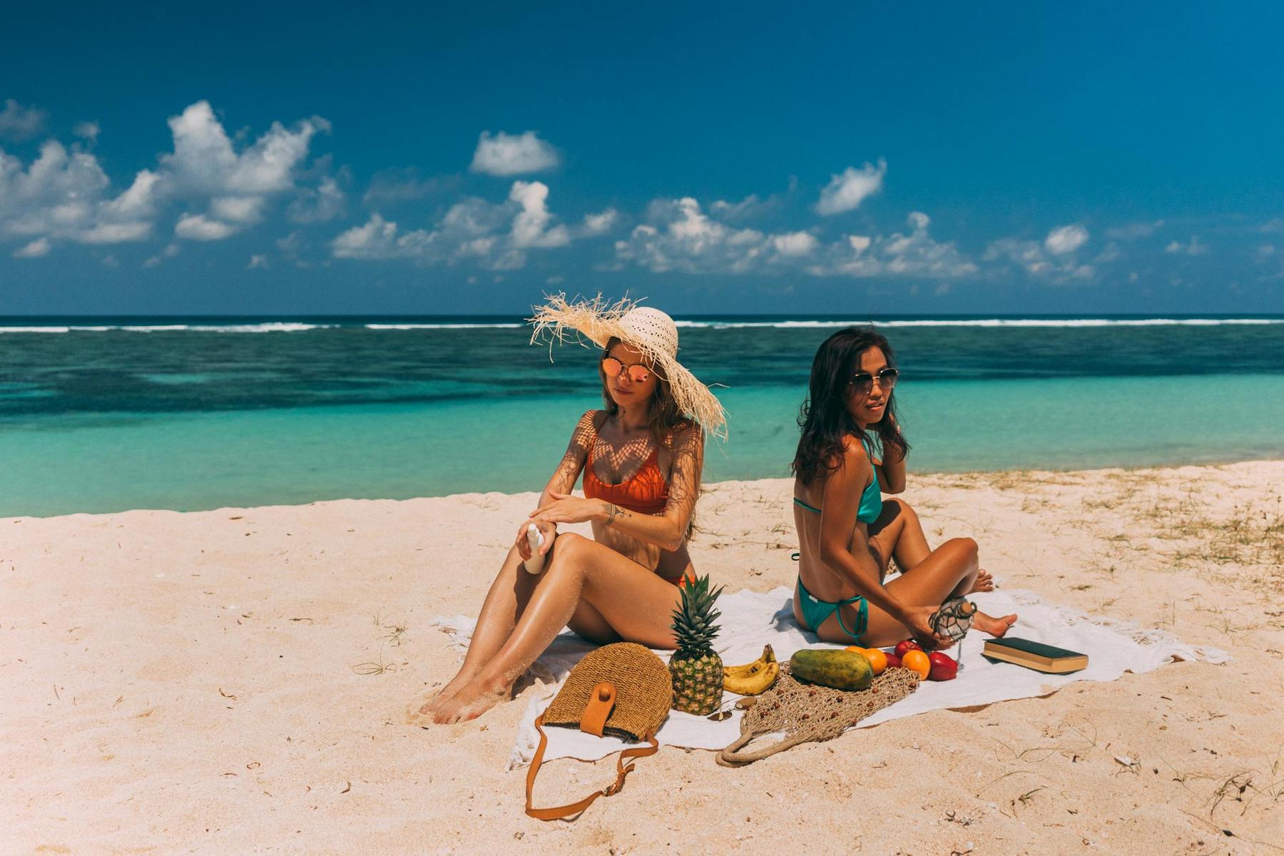 Two women in swimsuits sit on a beach towel by the ocean, surrounded by a hat, bag, fruit, and snacks on a sunny day.