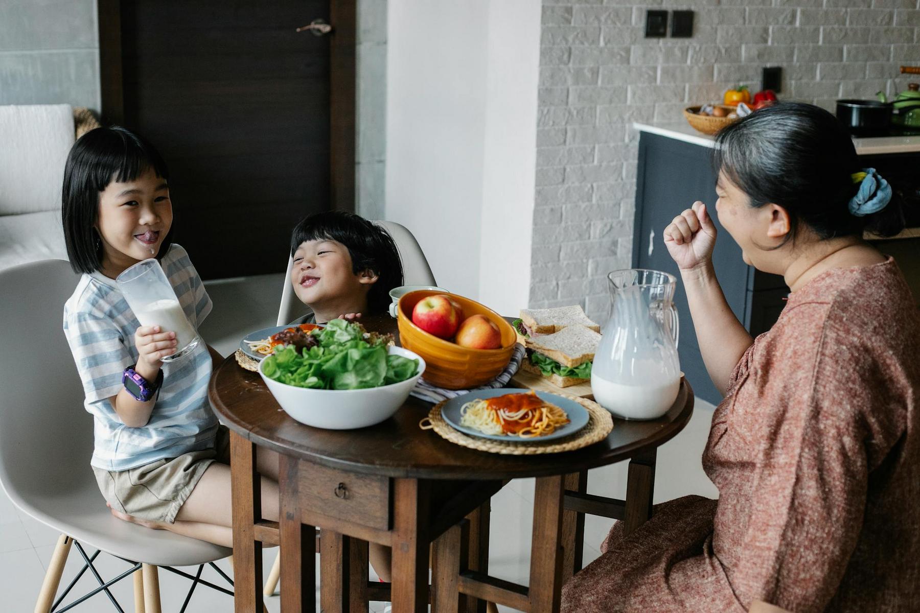 A woman and two children sit at a round table with food and drinks. The table has a bowl of salad, a plate with spaghetti, a bowl of fruit, a sandwich, and a pitcher of milk.