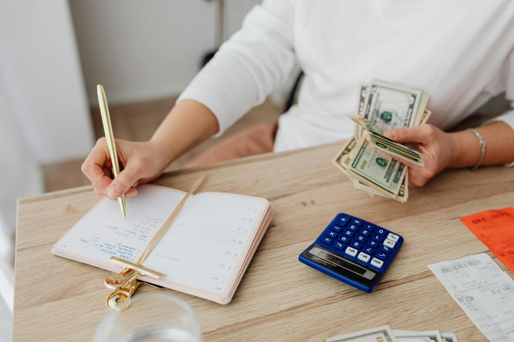 Person writing in a notebook while holding dollar bills, with a blue calculator and receipts on a wooden table.