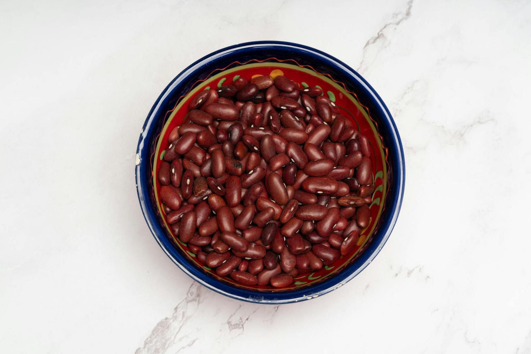 A colorful bowl filled with uncooked red kidney beans sits on a white marble surface.