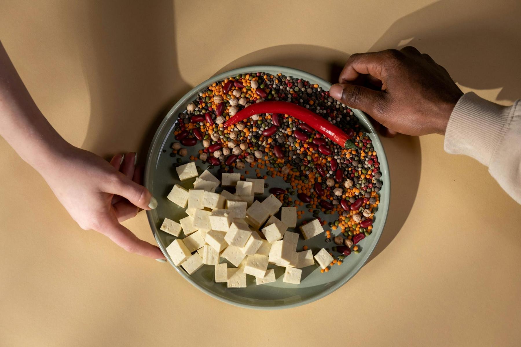 Two hands hold a plate with diced tofu, a variety of beans, lentils, peas, and a red chili pepper on a beige surface.