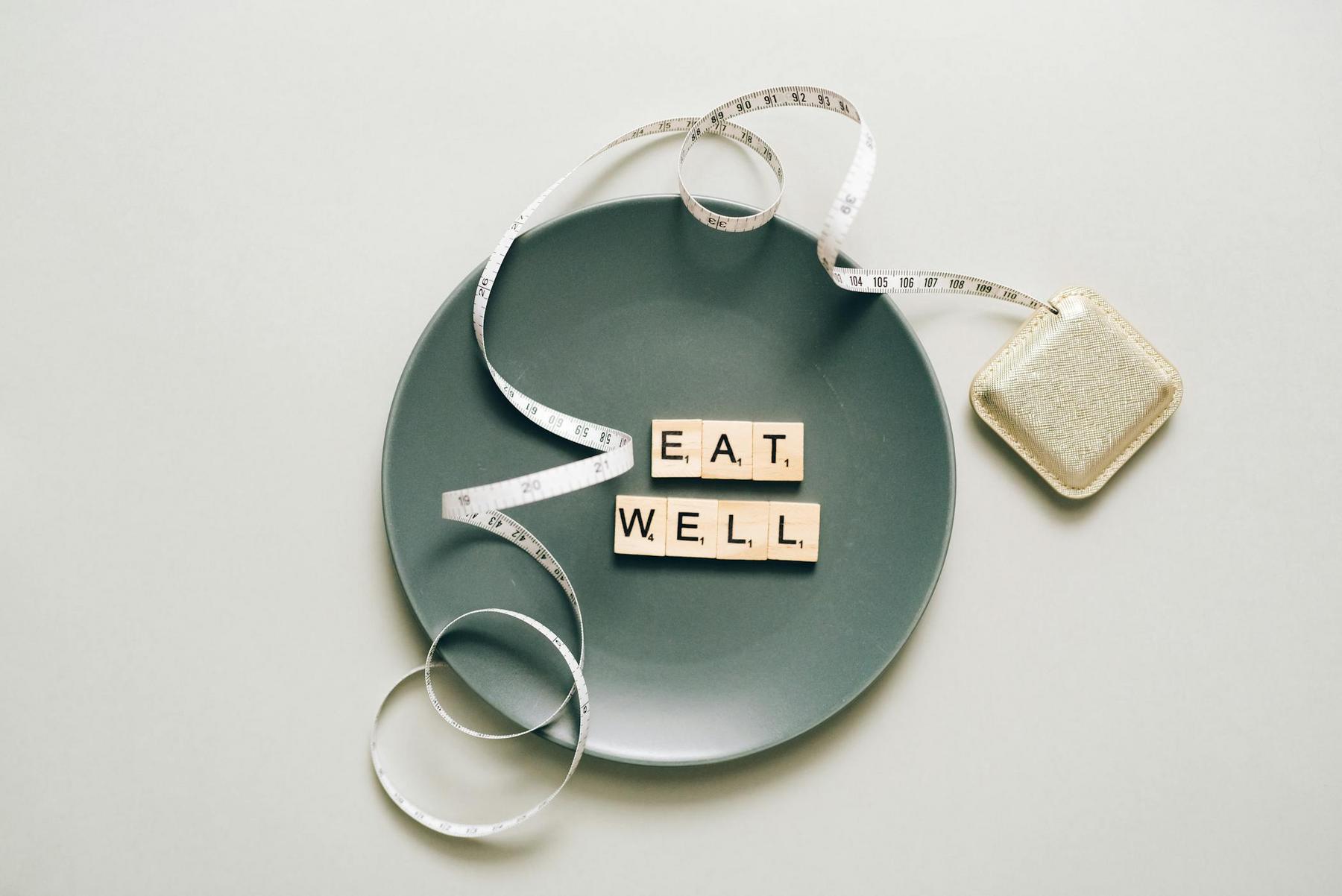 A gray plate with Scrabble tiles spelling "EAT WELL," surrounded by a coiled measuring tape, on a light surface.
