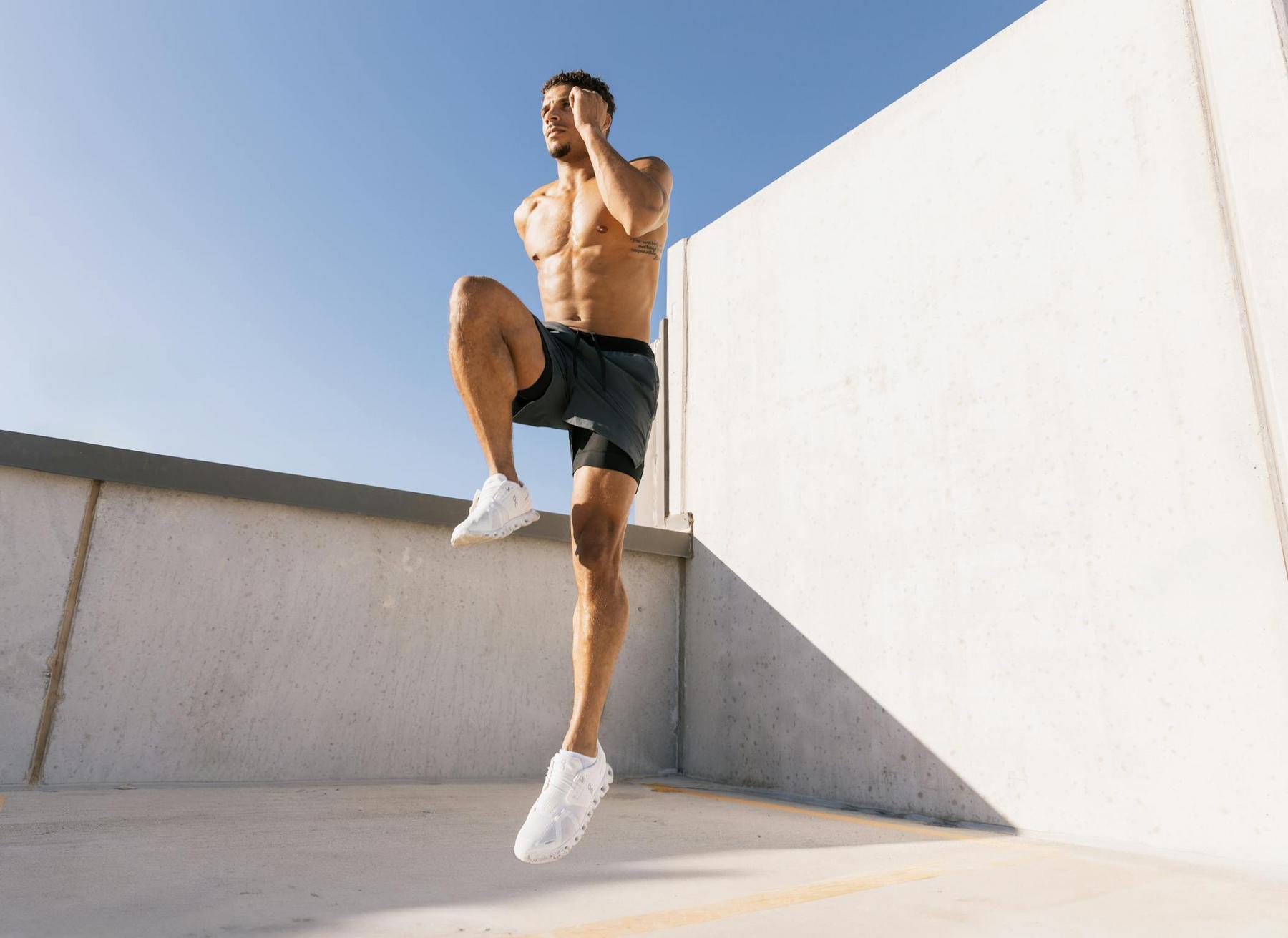 Shirtless man in black shorts and white sneakers does a high knee exercise outdoors against a concrete wall on a sunny day.