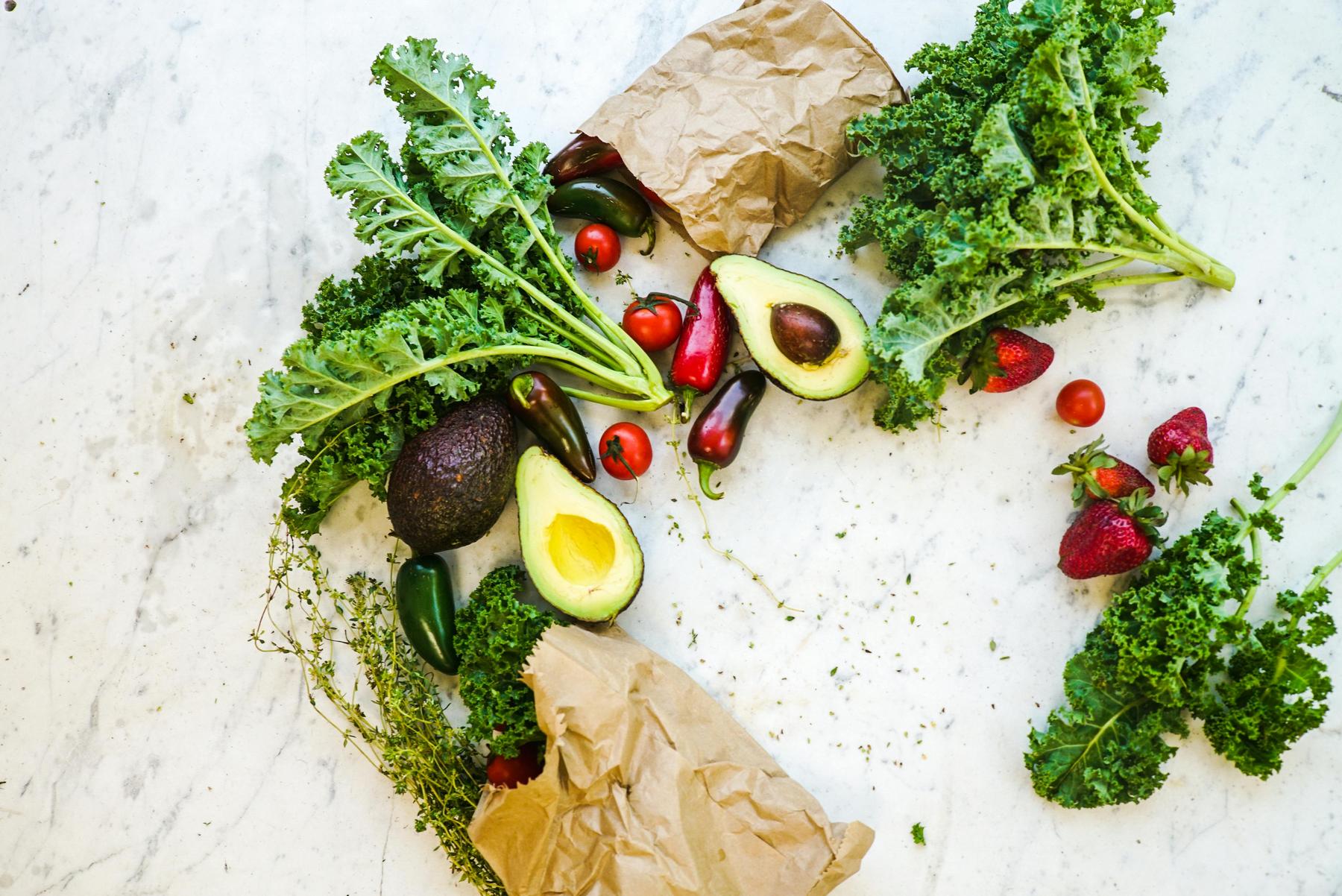 Avocado halves, kale, cherry tomatoes, strawberries, jalapeños, and herbs are spread on a marble surface with brown paper bags.