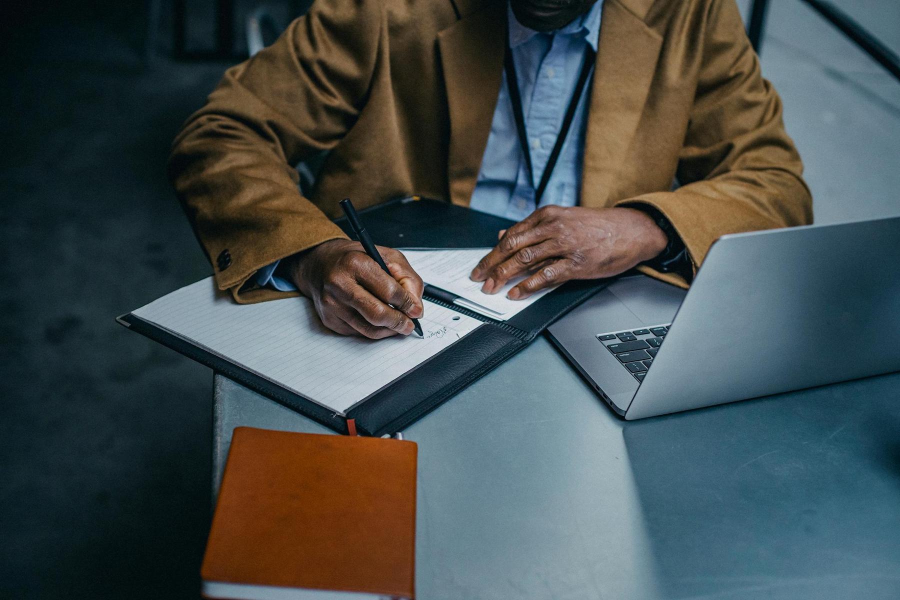 Person in a brown jacket writing in a notebook next to a laptop on a table.